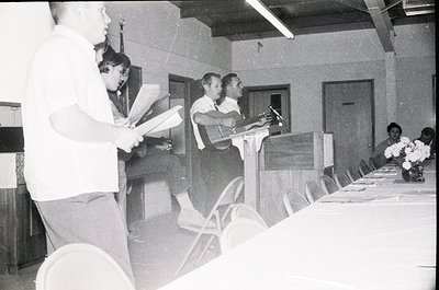 Black-and-white indoor shot of a formal event in the 1960s–70s, featuring a podium with a speaker addressing an audience. Mid...