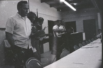 Black-and-white photo of a 1960s–1970s conference or meeting. Three individuals at a long table with microphones, suggesting ...