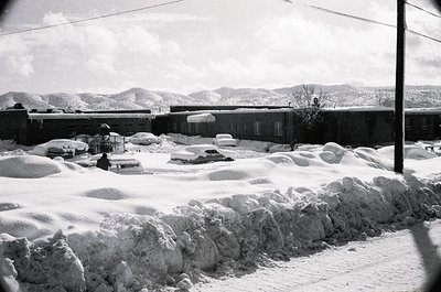Snow-covered urban street with buried vehicles, mid-20th century architecture. Heavy snowbanks line the roadside, partially o...