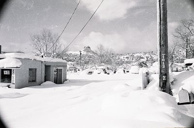 Snow-covered residential street with mid-century concrete block houses, blanketed in winter. Bare trees and utility poles lin...