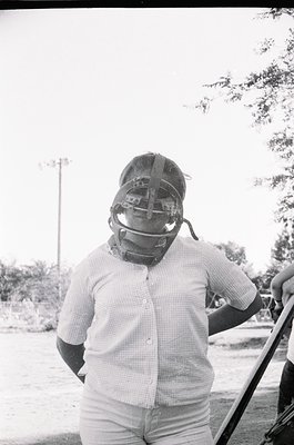 Baseball catcher in vintage protective gear, likely mid-20th century. Checkered shirt and knee-high socks suggest 1950s–1960s...