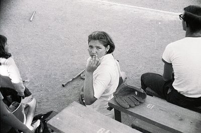 Baseball dugout scene featuring a woman in mid-1960s attire—short-sleeve blouse, skirt, and finger on lip—sitting beside a ca...