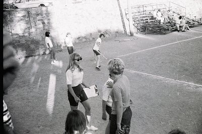 1970s outdoor basketball game in a concrete court setting. Players in short-sleeve shirts and shorts, with one holding a clip...