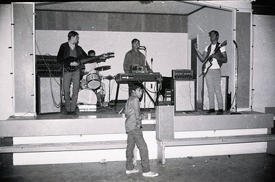 Vintage live band performance on a modest stage with four musicians: electric guitar, keyboard, drums, and bass. Child in for...