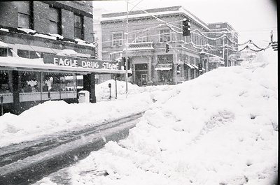 Mid-20th century street scene blanketed in snow, featuring the **Eagle Drug Store** with Art Deco signage. Snow-covered build...