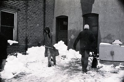 Two men clear snow from a brick-paved alleyway in mid-20th century, likely 1950s–1960s. One shovels snow into a pile, the oth...