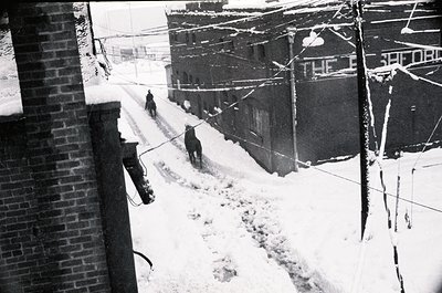 Mid-20th century urban alleyway blanketed in snow, with three workers clearing paths using shovels. Brick buildings flank the...
