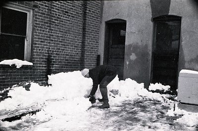 Mid-20th century black-and-white photo of a man clearing snow from a brick sidewalk. He wears a long coat, cap, and boots, sh...