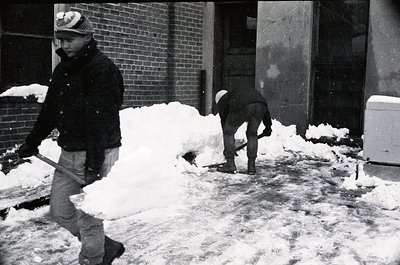 Two men clear snow from an urban sidewalk in mid-20th century, likely 1950s–1970s. One shovels while the other walks with a t...