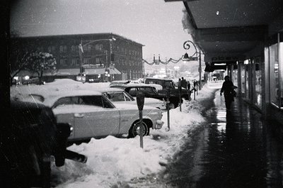 Vintage black-and-white street scene with mid-century cars parked along a snow-covered sidewalk. Classic 1950s–60s architectu...