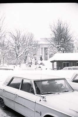 Snow-covered urban street with mid-century cars, likely 1960s–1970s. Prominent white sedan in foreground, leafless trees, and...