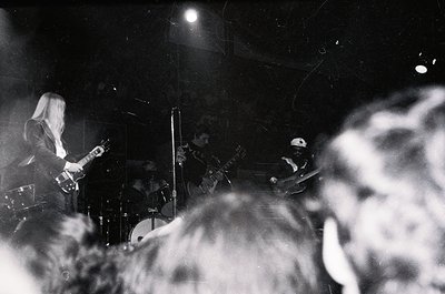 Three musicians performing live in dimly lit venue: guitarist (left), drummer (center), bassist (right). Crowd blurred in for...