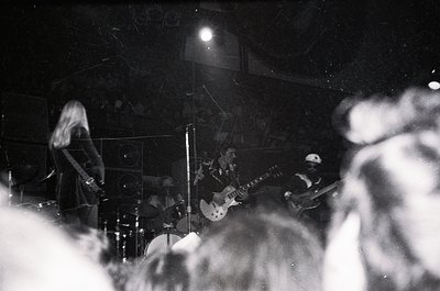 Intimate live band performance in dimly lit venue, featuring guitarist (left) and bassist (right) under spotlight. Crowd silh...
