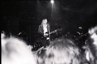 Guitarist performing live on stage, backlit by spotlight. Long hair and dark attire contrast against dark venue. Crowd silhou...