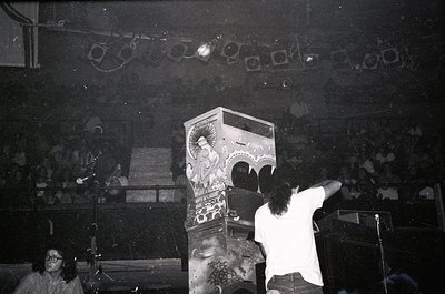 Vintage black-and-white concert shot featuring a DJ operating a large, retro arcade-style jukebox/music machine on stage. Cro...
