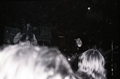 Vintage black-and-white shot of a live band performance in low-light, likely 1970s–1980s. Guitarist and drummer visible, with...