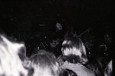 Black-and-white concert shot: stage lit by spotlight, guitarist playing electric guitar, drummer behind cymbals, audience sil...