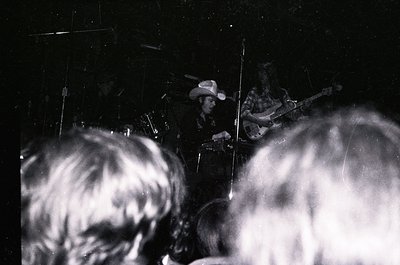 Blurred black-and-white concert shot: two musicians on stage—one playing drums (cowboy hat), one electric guitar—with audienc...
