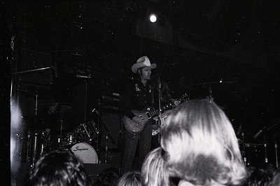 Black-and-white concert shot featuring a guitarist in cowboy hat playing a white electric guitar on stage. Stage lighting hig...
