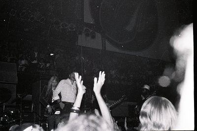Vintage black-and-white concert shot: performer on stage in checkered shirt, arms raised, with audience hands visible in fore...