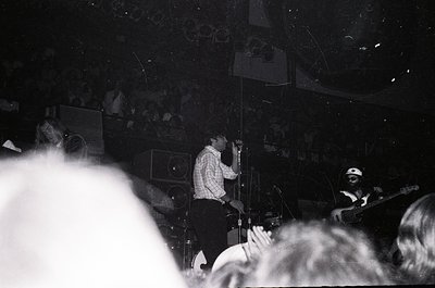 Black-and-white concert shot featuring a saxophonist and guitarist performing on stage, surrounded by a dense crowd. Stage li...