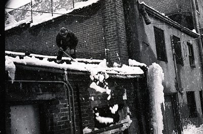 Black-and-white street scene: man clearing heavy snow from rooftop gutter using a shovel. Snow accumulates on brick buildings...