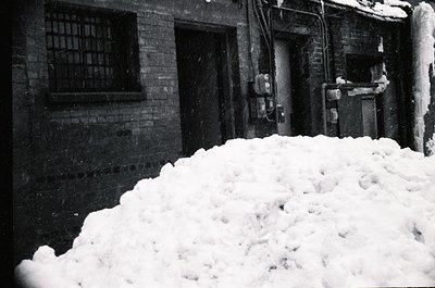 Mid-century industrial courtyard blanketed in deep snow, with brick walls and barred windows. Snowbanks obscure a fire escape...