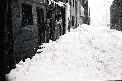 Snowdrifts pile high along a narrow alleyway flanked by brick buildings, likely residential or mixed-use. Heavy snowfall obsc...