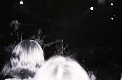 Black-and-white shot of a live jazz performance in a dimly lit venue. Backlit saxophonist playing center stage, with blurred ...