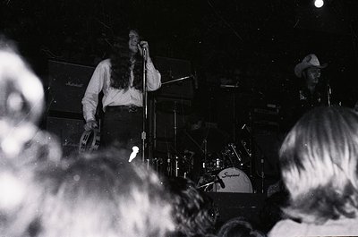 Vintage black-and-white concert shot of a band performing live, likely 1970s. Frontman in long hair and cowboy hat sings into...