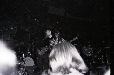 Black-and-white live band performance in dimly lit venue, featuring guitarist with checkered shirt and long hair playing elec...