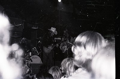 Vintage black-and-white concert shot featuring a guitarist in a cowboy hat playing electric guitar on stage. Crowd silhouette...