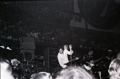 Vintage black-and-white concert shot of a guitarist performing onstage, framed by vintage amplifiers and a packed crowd. Stag...