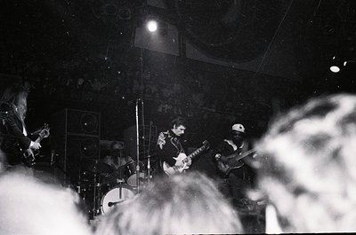 Vintage black-and-white live band performance in dimly lit venue, featuring guitarist, drummer, and vocalist. Stage lighting ...