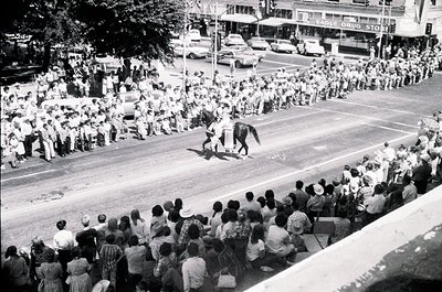 Aerial view of a mid-20th-century parade featuring a mounted police officer leading a crowd. Spectators line both sides of a ...