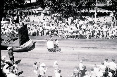 Aerial shot of a mid-20th-century outdoor event featuring a large crowd in casual summer attire. Central figure in a white su...