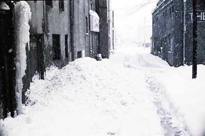 Blizzard-covered urban alleyway with heavy snow accumulation on buildings and sidewalks, likely mid-20th century. Snowdrifts ...