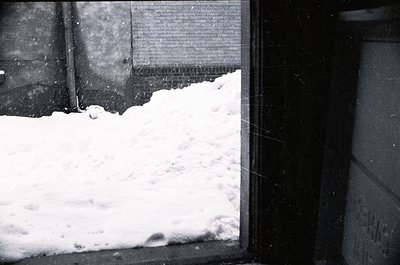 Black-and-white urban winter scene: snow piled against a brick wall and window frame, suggesting heavy snowfall. Industrial p...