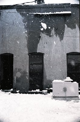 Snow-covered urban facade with three tall, narrow windows framed by worn concrete. Overhead wires and a utility box with snow...