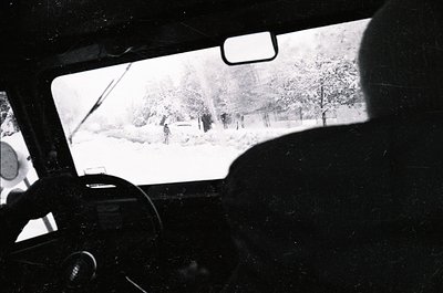 Black-and-white shot from inside a vintage car, snow-covered landscape visible through windshield. Distant figures in winter ...