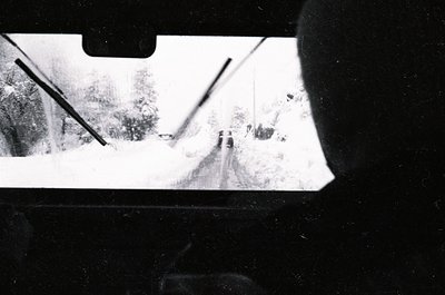 Blurred rearview shot of a snow-covered road with minimalist composition—bare trees, faint tire tracks, and a lone figure in ...