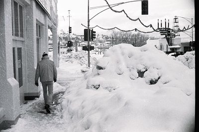 Black-and-white street scene featuring a lone man in winter attire navigating a snow-covered sidewalk. Snowbanks line the roa...