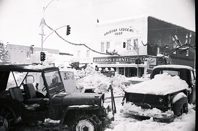 Mid-20th century snowstorm disrupts downtown streets, with deep drifts covering vehicles and sidewalks. Snow-laden traffic li...