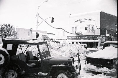 Mid-20th century street scene after heavy snowfall, featuring a tractor clearing snow in downtown. Visible: "Arizona Lodging"...