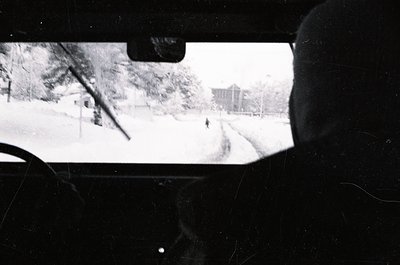 Black-and-white shot from inside a vehicle, framed by windshield and rearview mirror. Snow blankets a residential street with...