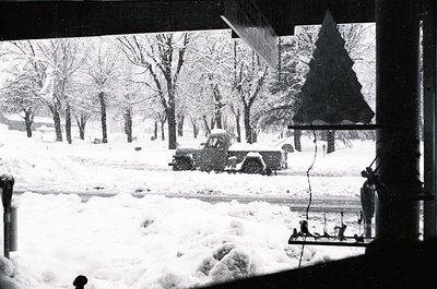 Vintage black-and-white winter scene showing a snow-covered urban park. Snow-laden vintage truck parked near leafless trees, ...