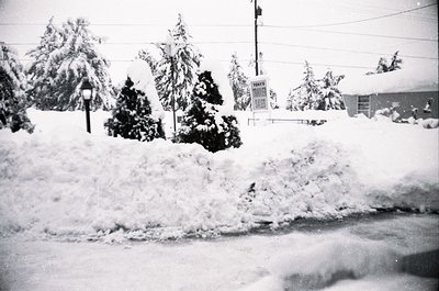 Black-and-white street scene blanketed in heavy snow, likely mid-20th century. Snow-covered evergreens, utility poles, and a ...