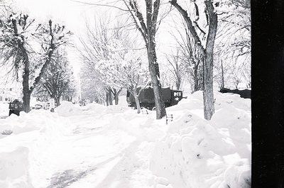 Mid-20th century winter street scene with deep snow covering a residential road. Bare trees line both sides, and a vintage ca...