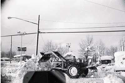 Mid-20th century winter street scene with heavy snow accumulation. Snow-covered utility truck and parked cars on a residentia...