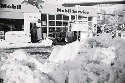 Mid-20th century Mobil Service station buried under heavy snowfall. Two workers clear snow with a snowblower and shovel near ...